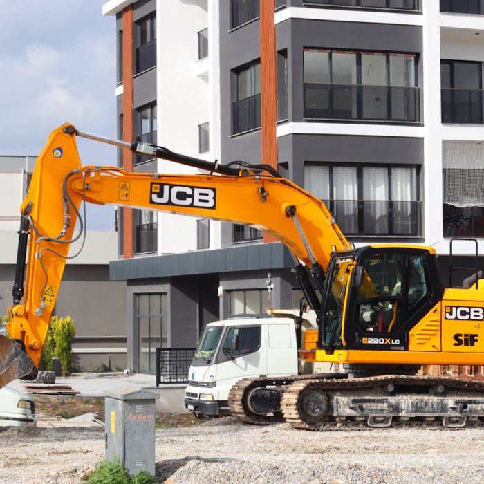 A yellow excavator operating at a city construction site in front of modern buildings.